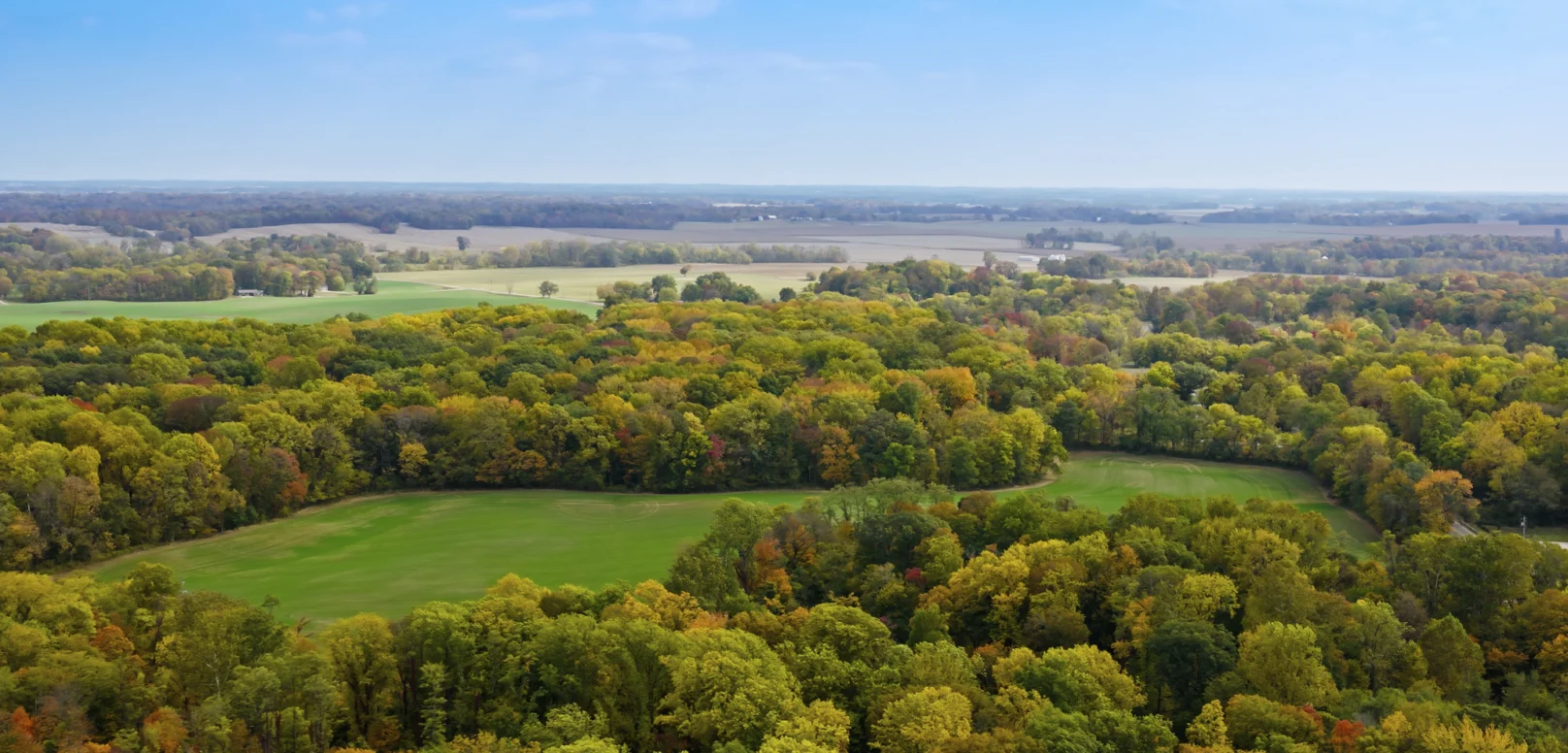 Aerial view of a landscape with patches of green fields surrounded by autumn-colored forests under a clear blue sky.