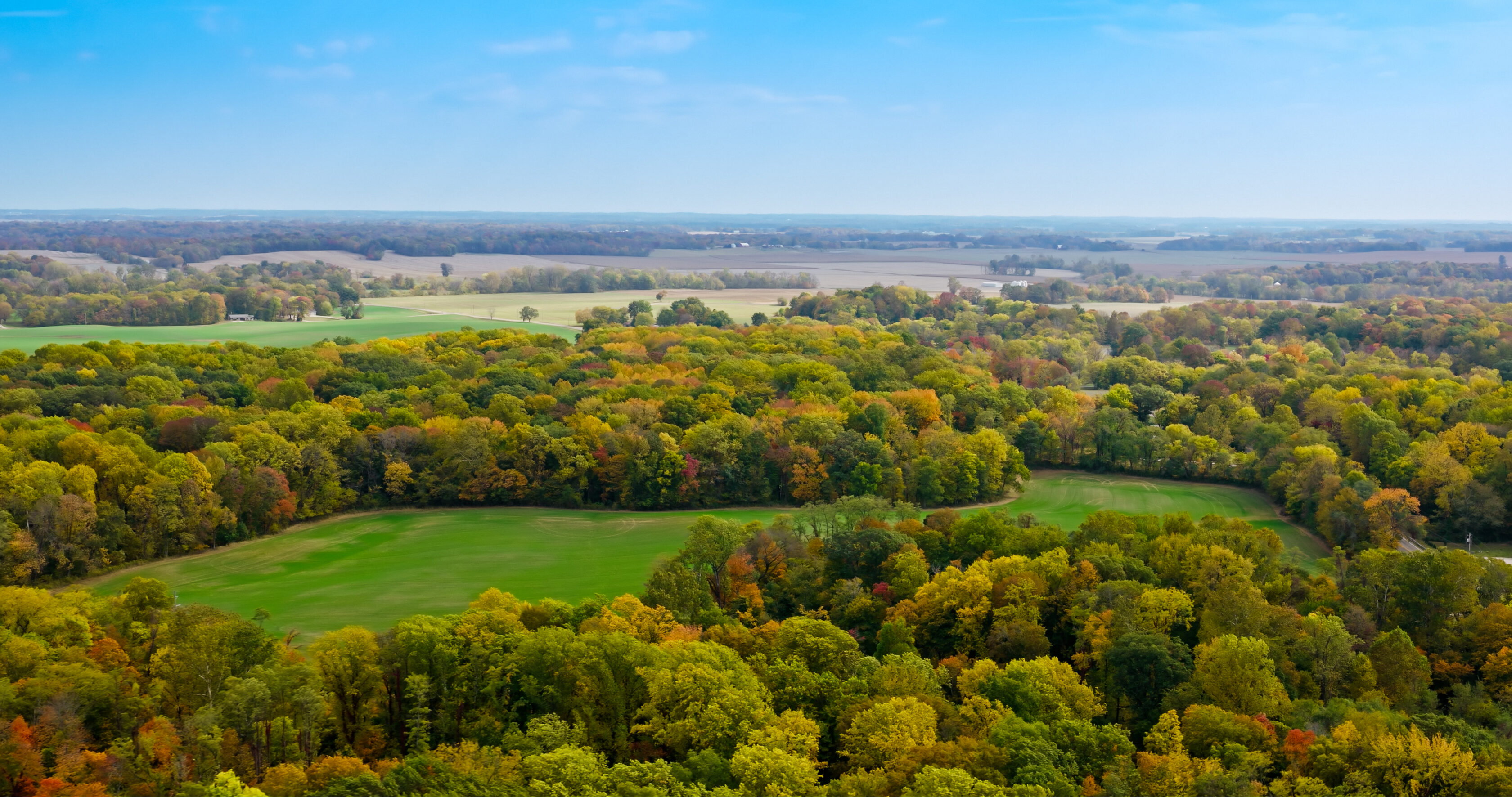 An aerial view of a landscape featuring patches of green fields surrounded by autumn forests in shades of green, yellow, and orange.